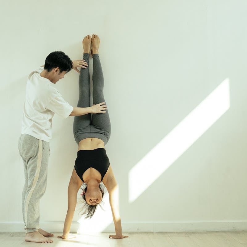 Full body of unrecognizable male instructor helping young female in activewear doing handstand in light room